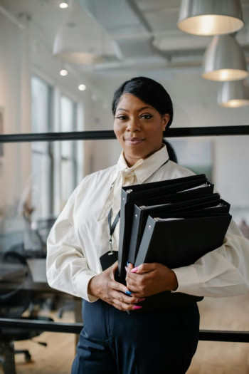 A confident black woman stands holding files. She has long black hair in a low ponytail and is wearing a white shirt and dark blue jeans.