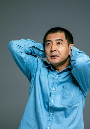 A mature Asian man with dark hair and a blue shirt stands looking up with his hands behind his head