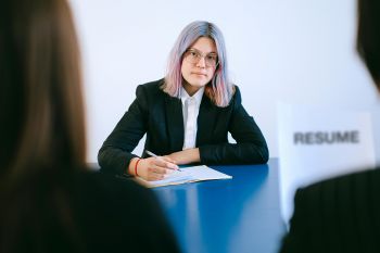 A woman with pink and blue hair sits listening to two others. She has glasses and holds a pen over a notebook.