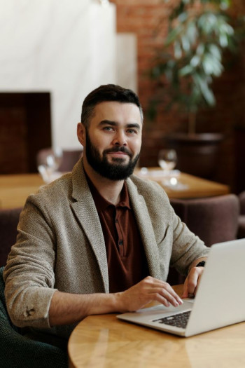 A bearded man with dark hair sits at a table at an open laptop. He is looking at the camera and looks kind. He is wearing a beige blazer and a dark red-brown polo top.