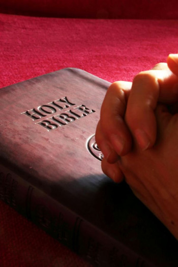 Two hands clasped in prayer on top of a Bible