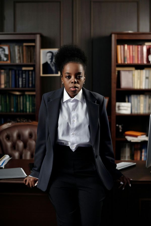 A black business woman leans against a desk in a lawyer's office. She has black hair tied up and is wearing a black suit with white shirt.