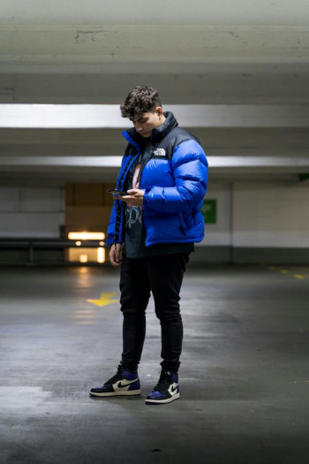 A man in his 20s stands in a car park looking at a phone. He is wearing a blue puffer jacket and has tight black jeans and blue sneakers. He has short curly hair and is white.