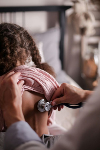 A doctor or nurse listens to the back of a girl. She has a pink top on and brown curly plaited hair. She is facing away.