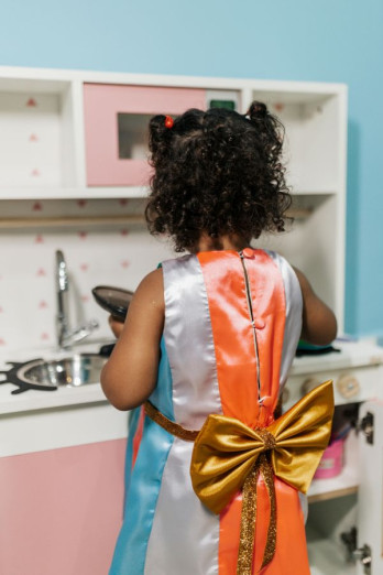A small girl is dressed in costume and playing in a toy kitchen. She is facing away from the camera. 