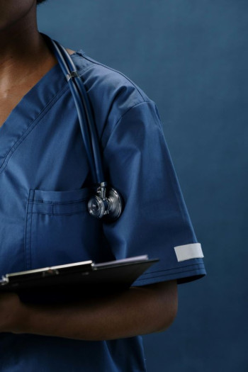 A close crop of a nurse's left shoulder showing them wearing scrubs and holding a clipboard.