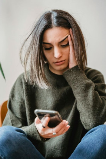 A white woman with brown hair looks down at her phone in a concerned way. She has a green jersey and blue jeans on.