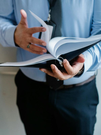 A close up shows a man wearing a blue shirt, blue pants, and a tie. He is flipping through a book that looks like a corporate diary.
