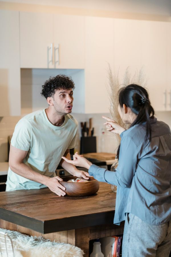 A couple are fighting and there is a table between them. The man has shirt, dark, curly hair and a light t-shirt. The woman is wearing grey pants and a grey cardigan. She has dark hair in a ponytail.