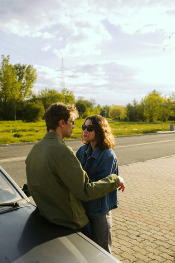 A man in a green jacket with short dark hair and sunglasses leans backwards onto a car. A woman stands closely in front of him. She has shoulder length dark wavy hair, sunglasses and a denim jacket.