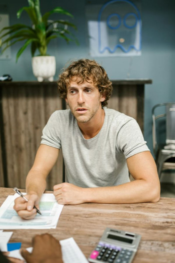 A man with sandy curly hair and a beige t-shirt sits at a desk with a pen in his hand.