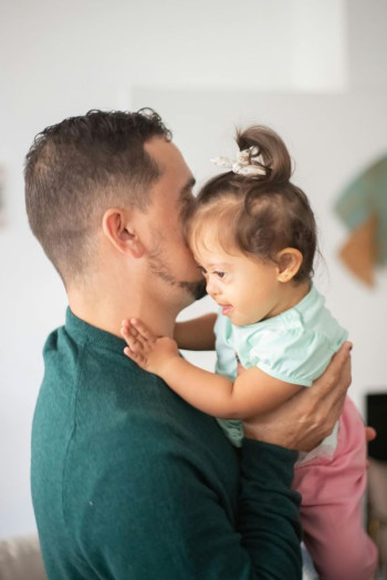A baby is held by a man wearing a green shirt who has very short dark hair. We can't see his face. The baby is a girl in a mint top and pink pants and has Down's Syndrome.