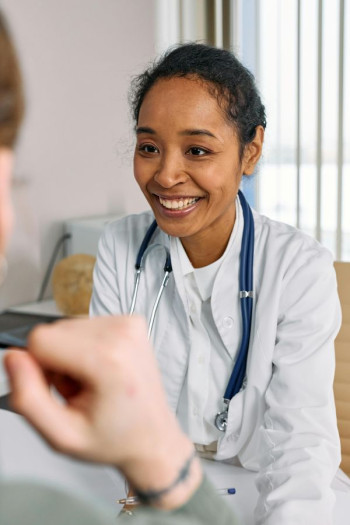 A black woman doctor in a white coat sits speaking to another adult.