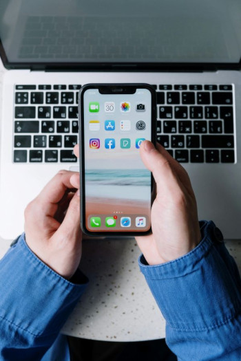 A pair of white hands holds a smart phone over top of a laptop keyboard. The person wears a blue shirt.