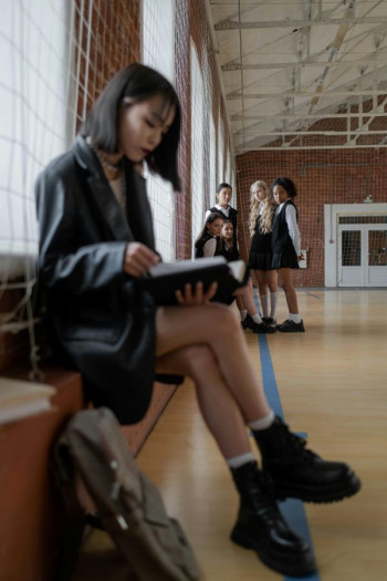 A high school girl sits on a bench in the foreground wearing a short skirt and black blazer. She has a file open looking at it. In the background three teenage girls are standing talking.