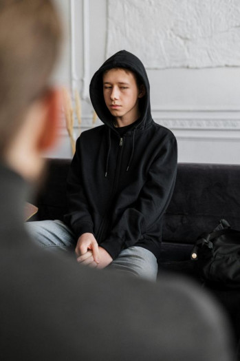 A sad looking teenager sits on a couch wearing a black hoody. An adult sits opposite him.