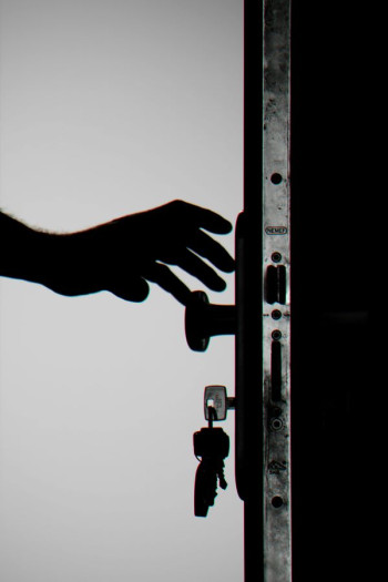 A black and white image shows the silhouette of a person's hard reaching to open a door that is already slightly open. We can see the keys in the lock too.