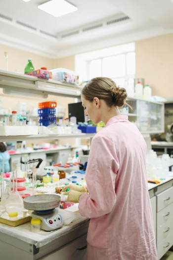 A lab worker faces away from the camera in her lab. She has a pink lab coat and has light brown hair in a low bun.