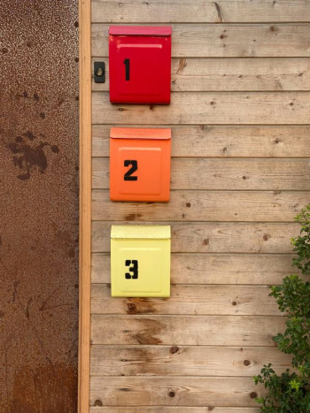 Three mailboxes on a wooden wall. The top one (1) is red, middle (2) is orange, and bottom (3) is lemon yellow.
