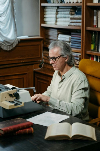 A man with shoulder length grey hair and round glasses sits at a word processor. He is wearing a beige shirt and surrounded by shelves of books.