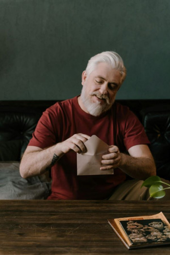 A man with short white hair and beard opens an envelope. He is wearing a maroon tshirt.