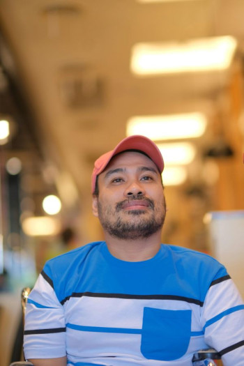 A smiling man sits looking happy. He is wearing a red cap and t-shirt with blue and white stripes. 
