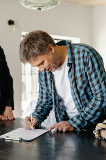A man with short grey hair leans over a table signing a document. He is wearing an open checked shirt with a white tshirt under it. 