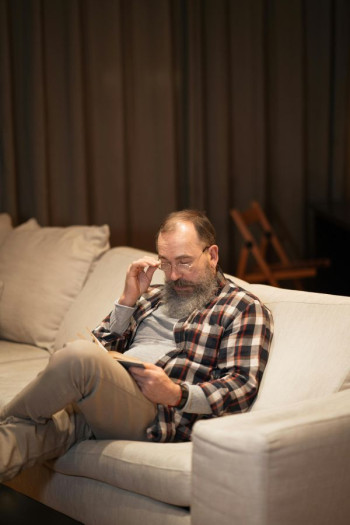A bearded white man with thinning hair sits on a couch. He is wearing glasses, touching them as he looks at a book. He is wearing a black, white, and red check shirt and beige pants.