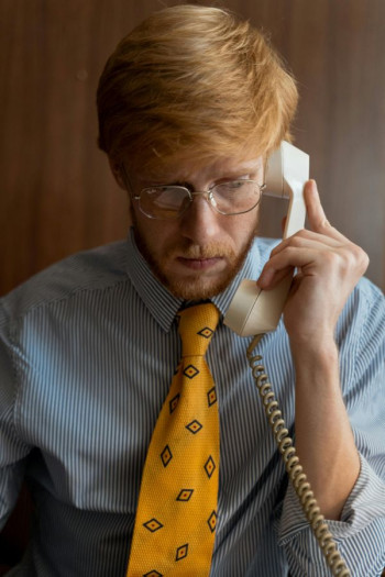 A man wearing a blue striped business shirt and yellow tie speaks on a landline. He has short strawberry blond hair and thin wire framed glasses.