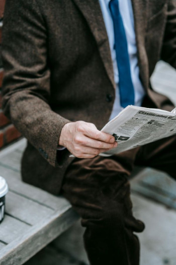 A man in a brown suit and blue tie and shirt sits on a bench reading the newsletter. We can not see his face.