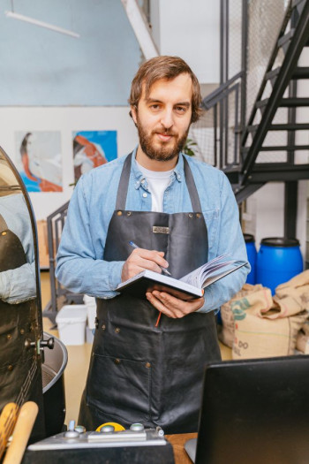 A man in an apron stands in a storeroom holding a pen and journal