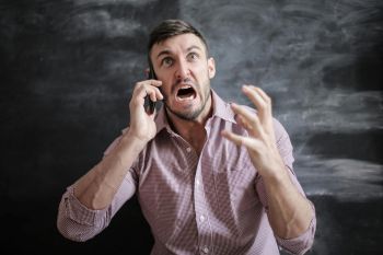 A man in a pink shirt is yelling into a phone. He is white with dark short hair and a goatee.