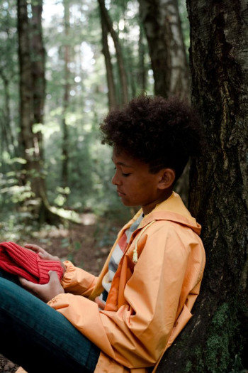 A boy in an orange jacket holding a red hat leans against a tree in a forest.