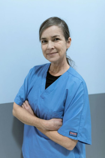 A nurse in blue scrubs stands side-on with her arms folding looking at the camera. She has a grey ponytail and is smiling.