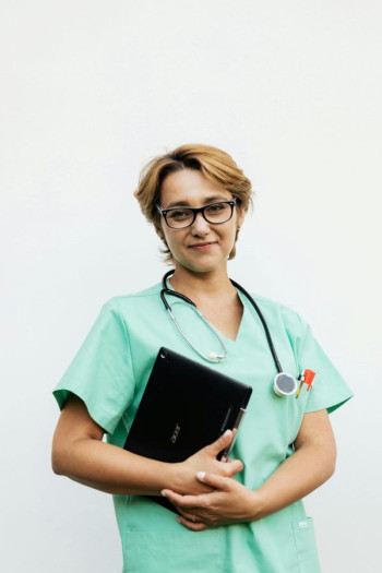 A nurse in green scrubs stands holding a folder or iPad. She has short dark blonde hair and glasses.