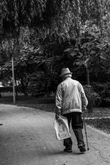 An old man walks away from a dirt road. He is wearing a hat, jacket, dark pants and has a walking stick. He is also carrying a plastic bag.