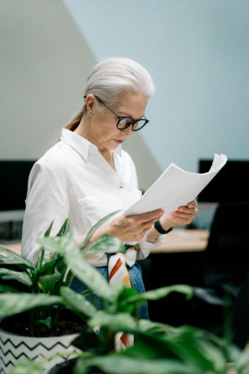 An older woman with a grey ponytail and black glasses looks at a piece of paper she is holding. She is wearing a white shirt.