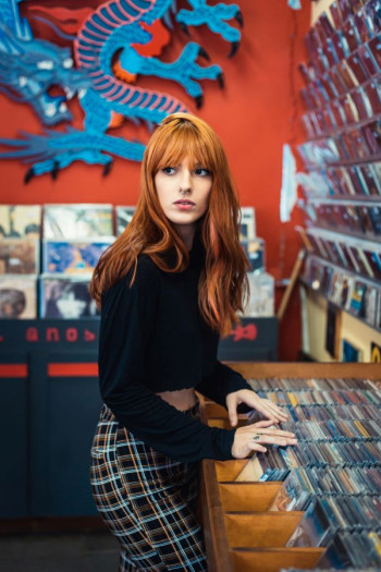 A woman with long red hair and a fringe stands sideways looking behind her. She has her hands in a bin of CDs. She is white and wearing a plain black top and tartan pants.