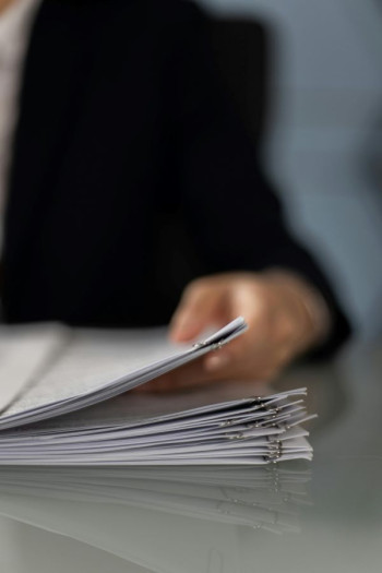 A person's hand lifts a report off a stack of reports on a desk.