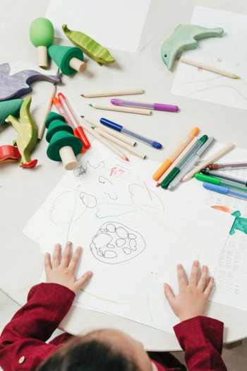 A child's hands rest on a table that is covered with a drawing and pens.