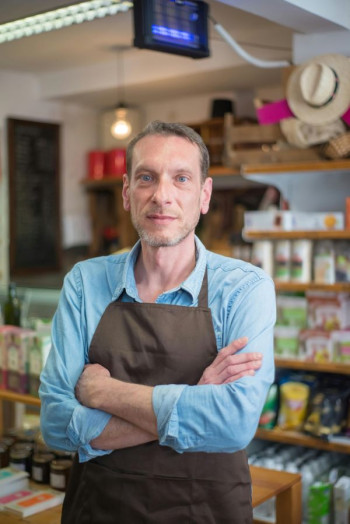 A white man wearing a blue long sleeved shirt and brown apron stands with his arms folded looking at the camera. He is in a shop.