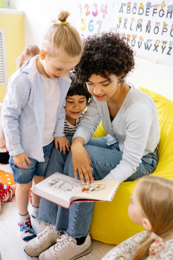 A woman with dark curly hair sits on a yellow chair reading a story to three or four children.