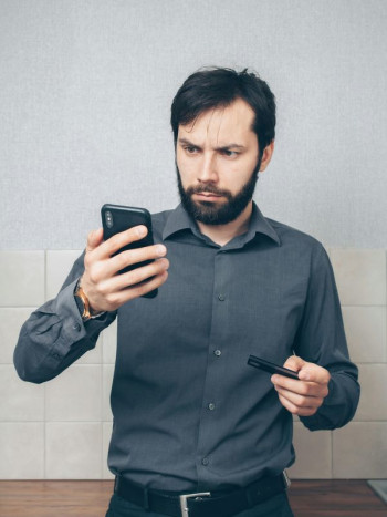 A white man with black hair and beard looks at his phone. He is perplexed. He wears a dark grey shirt and dark pants.