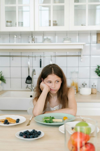 An uphappy child sits at a table with plates of fruit and pancakes. She is white with long, dark brown, straight hair.