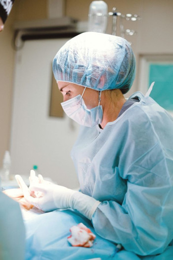 A female surgeon is at the operating table wearing full blue scrubs and face mask.