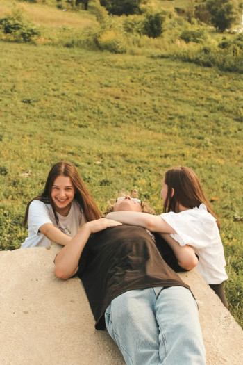 Three teenagers hang out outside. Two are girls wearing white t-shirts who have long brown hair. The other is a boy lying down wearing a black top and faded jeans.