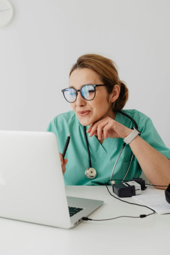 A doctor in a scrub top looks at a computer screen. She is animated. She has light brown hair in a ponytail and has glasses.