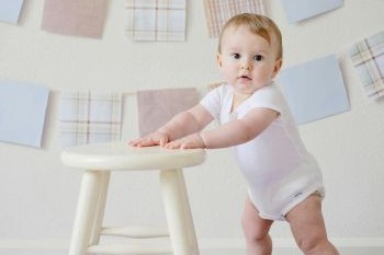 A white toddler stands holding onto a cream stool
