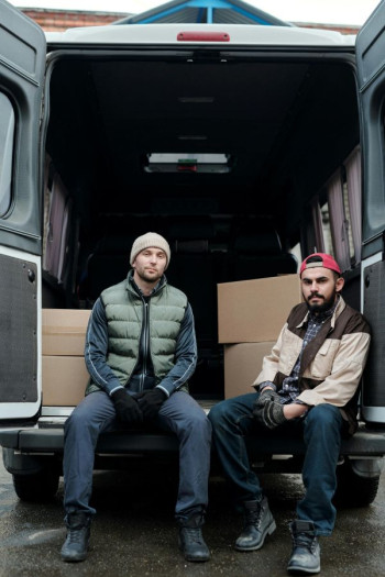 Two men wearing hats, jackets, and jeans sit casually in the back of an open van. There are boxes in the van.