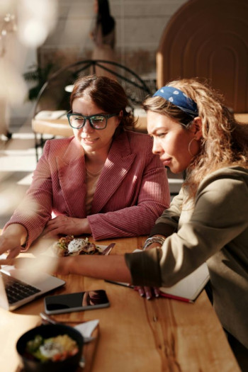 Two women sit at a table looking at a shared laptop. The woman on the left has a pink blazer, brown bobbed hair, and glasses. The woman on the right has curly blonde hair and a headband that is blue. She is wearing a beige blazer.